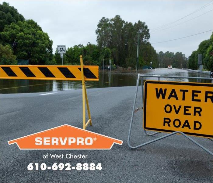 Road barriers and a sign reading “Water Over Road” are posted in front of a flooded street.